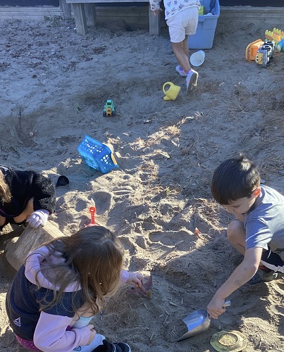 4 School age children in the sandbox, 3 are digging in the sand, one is walking away