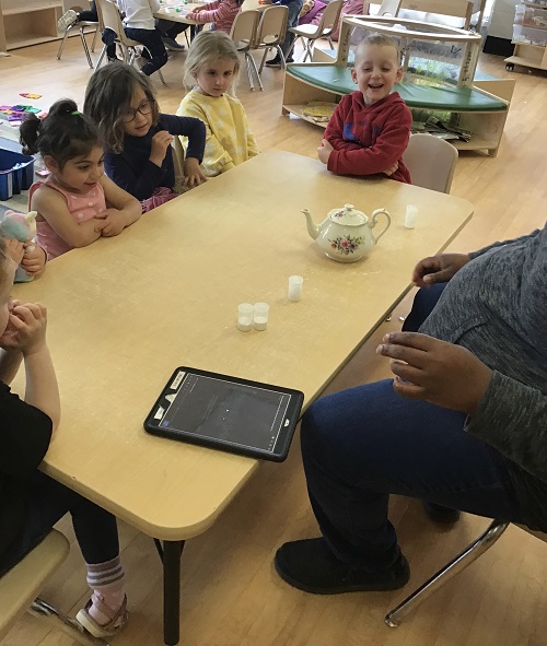 four children sitting across from an educator at a table. on the table there is a tea pot and 4 small clear cylyndrical containers and an ipad.