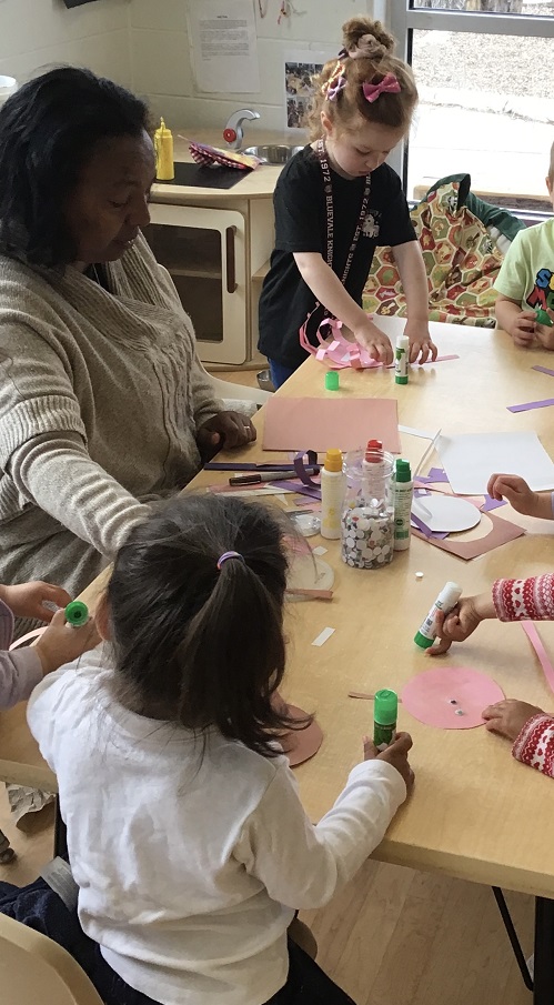 educator sitting at a table with children engaing in a craft with paper, glue sticks, googly eyes and bingo dappers