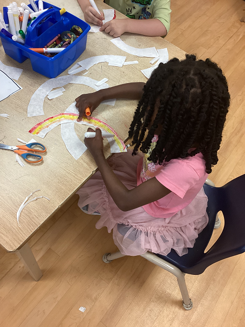 girl sitting at a table using an orange marker as she colours a rainbow