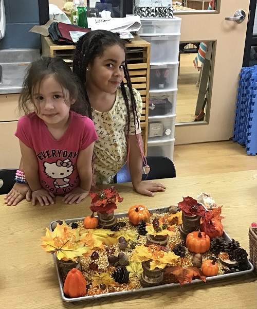 two children standing in front of a table. on the table is a tray full of artificial leaves, pinecones, acorns and popcorn seeds
