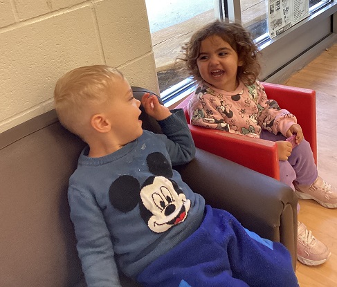 two children sitting in chairs one red one brown looking at each other and smiling/laughing