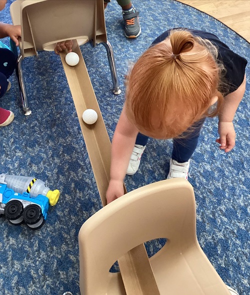 Child bending down placing a white ball down a ramp propped up on 2 chairs