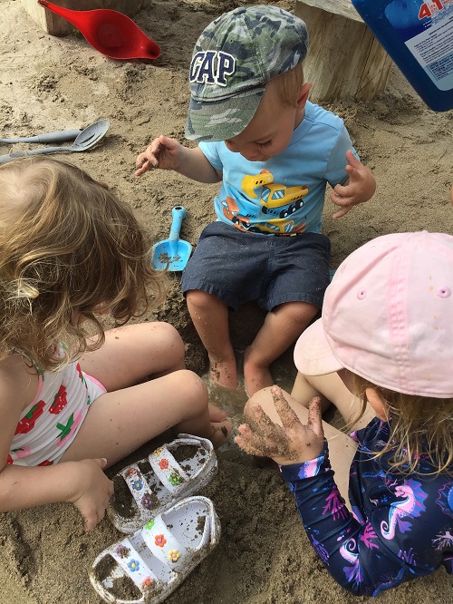 three children sitting with their feet in a puddle of water inside of the sandbox