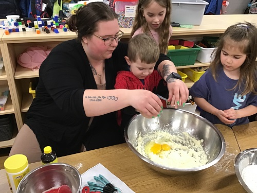 An educator helping a child crack an egg into a bowl of ingredients