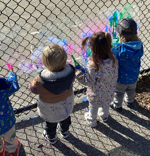 Children standing at a fence spraying paint onto a clear piece of plexiglass