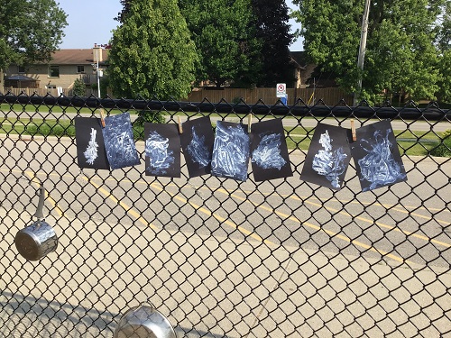 Multiple pieces of black construction paper painted with white sunscreen hanging on a fence in the sunlight