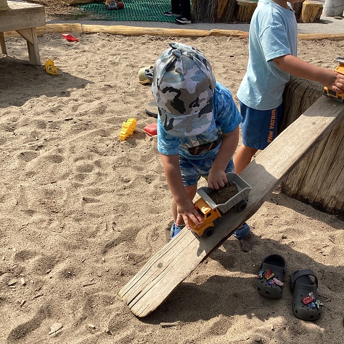 A child in the sandbox driving a truck down a plank of wood