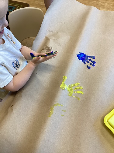 A child at a table rubbing her hands together with different coloured paint on them