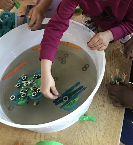 Bowl with water, popsicle sticks taped together with a child placing washers on the popsicle sticks - popsicle sticks floating around in the water with another child's hands hovering by