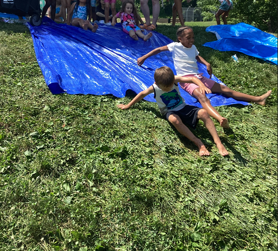 2 summer camp children sliding down a blue tarp smiling, one summer camp child sliding down behind with a handful of children standing at the top of the hill waiting their turn