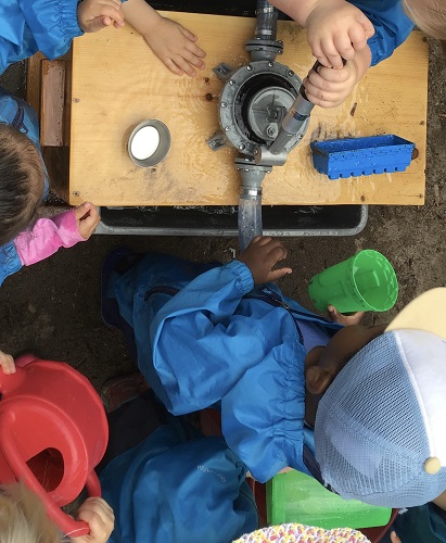 3 Toddler children around the water pump, one Toddler is using their hands and arms to make the pump spew out water for the other children