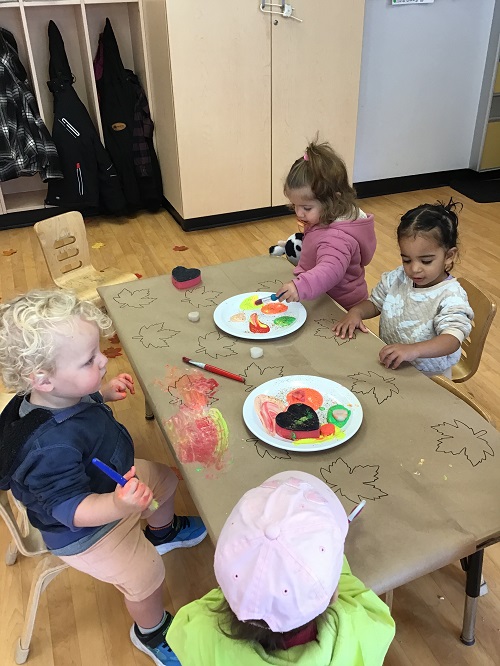4 Toddler children around the table with paint brishes and sponges and paint with paper with outlined leaves