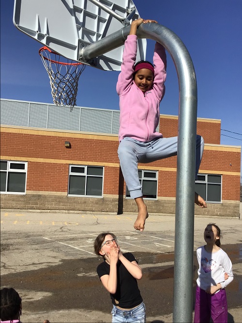 Child hanging off top of basketball net, while peers admire and encourage. 