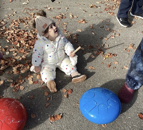 An infant using a metal drum and wooden drumstick outside