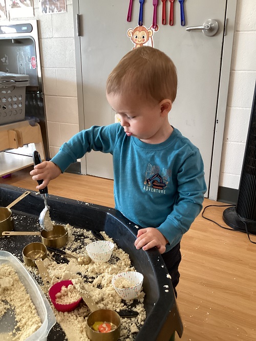 A toddler using a spoon to scoop up an oil and flour mixture