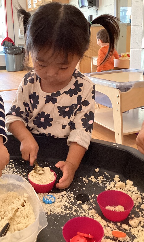 A toddler flattening an oil and flour mixture with a spoon inside a silicone muffin tray