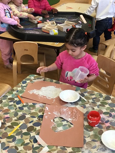 3 preschool children sitting in the background at the tuff tray with some blocks, chalk, and popsicle sticks. Preschool child sitting at the table creating with glue and sand